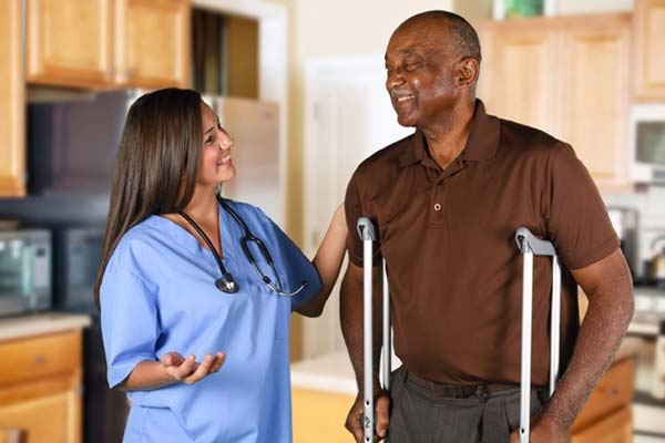 woman dressed in blue scrubs helping an elderly man on crutches